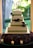 A beautifully decorated four-tiered wedding cake with intricate white icing patterns and green ribbons. It features a metal heart-shaped cake topper. The base is surrounded by purple flowers and lit candles, creating an elegant and romantic atmosphere with a lush garden visible through the window behind.