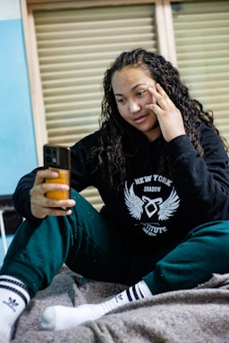 A person with long curly hair is sitting on a bed or couch while holding a smartphone. They are dressed casually in a dark hoodie and sweatpants, with socks featuring a logo. The background shows a partially open window with blinds.