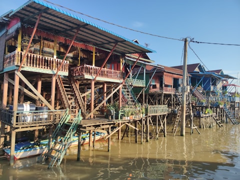 Several colorful stilt houses are built along a waterway, with wooden staircases leading up to the structures. The stilts are visible beneath the houses, elevating them above the water level. Vegetation and potted plants decorate the balconies, and there are various items stored beneath the houses. The scene is lit by sunlight, casting reflections in the water.