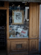 A shop window display featuring various items such as framed artwork, mugs, and bags with the word 'MERDE' prominently displayed. A sign reads 'JE SOUTIENS LA GREVE' indicating support for a strike. The wooden window frame suggests a rustic or vintage style.