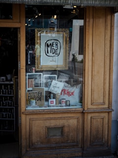 A shop window display featuring various items such as framed artwork, mugs, and bags with the word 'MERDE' prominently displayed. A sign reads 'JE SOUTIENS LA GREVE' indicating support for a strike. The wooden window frame suggests a rustic or vintage style.