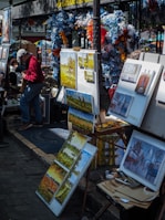 A street art market featuring various paintings on display, with colorful flowers and decorations around the stall. Several canvases depict landscapes and buildings, and there is an artist adjusting paintings on an easel.