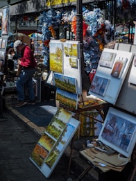 A street art market featuring various paintings on display, with colorful flowers and decorations around the stall. Several canvases depict landscapes and buildings, and there is an artist adjusting paintings on an easel.