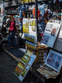 A street art market featuring various paintings on display, with colorful flowers and decorations around the stall. Several canvases depict landscapes and buildings, and there is an artist adjusting paintings on an easel.