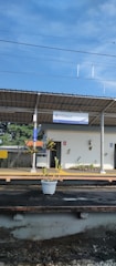 A small train station platform with a sign that reads 'Cilebut'. There is a single plant in a white pot on the platform. The platform is covered by a roof. The sky is clear and blue, and there are trees in the background.