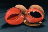 A vibrant red fruit is sliced open, revealing its juicy interior and a prominent black seed. The fruit has a rough brown exterior. The setting appears to be on a dark surface, possibly a table or counter, with a piece of wood partly visible underneath the fruit.
