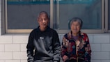 An elderly couple is seated side by side on a tiled bench outdoors, with a large window behind them. The man is wearing a black sweater with the word 'SPORT' printed on it, while the woman wears a colorful jacket with intricate patterns. Both appear content and relaxed, basking in the daylight.