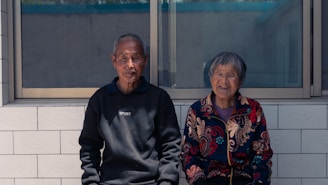 An elderly man and woman doing seated stretches together in a sunny community room.