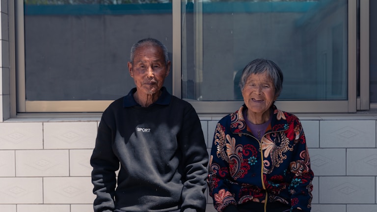 An elderly couple is seated side by side on a tiled bench outdoors, with a large window behind them. The man is wearing a black sweater with the word 'SPORT' printed on it, while the woman wears a colorful jacket with intricate patterns. Both appear content and relaxed, basking in the daylight.