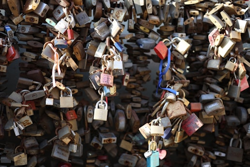 A dense collection of variously shaped and sized padlocks attached to metal chains. Some padlocks are rusty or weathered, while others have colorful finishes such as red, blue, and silver. The chains form a complex and intertwined pattern against a dark background.