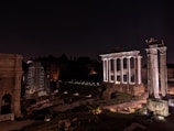 Ancient Roman ruins are illuminated at night, featuring tall, white columns and stone structures set against a dark sky. Some areas are under scaffolding, indicating restoration work.