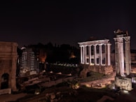 Ancient Roman ruins are illuminated at night, featuring tall, white columns and stone structures set against a dark sky. Some areas are under scaffolding, indicating restoration work.