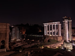 Ancient Roman ruins are illuminated at night, featuring tall, white columns and stone structures set against a dark sky. Some areas are under scaffolding, indicating restoration work.