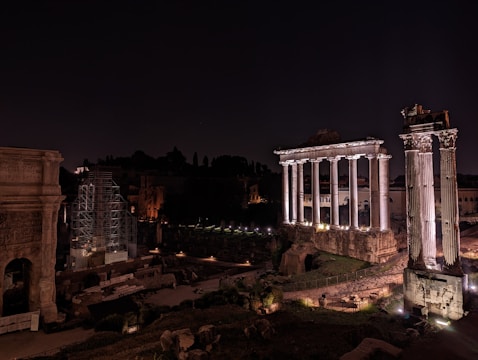Ancient Roman ruins are illuminated at night, featuring tall, white columns and stone structures set against a dark sky. Some areas are under scaffolding, indicating restoration work.