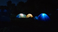 Three illuminated tents are set up in a dark outdoor setting, surrounded by trees. The tents have a glow from within, creating a cozy atmosphere in the otherwise dark environment.