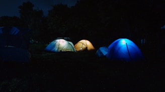 Three illuminated tents are set up in a dark outdoor setting, surrounded by trees. The tents have a glow from within, creating a cozy atmosphere in the otherwise dark environment.