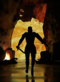 A silhouette of a warrior holding a sword in each hand, standing against a backdrop of fiery flames and rugged cave walls. The scene is dark, with glowing light in the background highlighting the warrior's form.