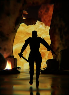 A silhouette of a warrior holding a sword in each hand, standing against a backdrop of fiery flames and rugged cave walls. The scene is dark, with glowing light in the background highlighting the warrior's form.