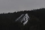 A dark, moody landscape featuring a forested mountain with a rocky cliff. Water trickles down the cliff, creating a thin waterfall. Overcast skies enhance the mysterious ambiance.
