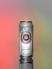 A tall, silver can with gold accents is positioned upright on a reflective surface. The label displays text in an ornate font with an emblem, indicating that the beverage is non-alcoholic and imported from Germany. The background is a smooth gradient transitioning from pink to gray and green hues.