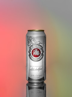 A tall, silver can with gold accents is positioned upright on a reflective surface. The label displays text in an ornate font with an emblem, indicating that the beverage is non-alcoholic and imported from Germany. The background is a smooth gradient transitioning from pink to gray and green hues.