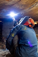 A welder wearing protective gear is working in a confined space, possibly under a structure. The bright arc of the welding torch illuminates the area, casting a blue and white light. Sparks fly as the metal is being welded, and the worker is crouched down, focusing intently on the task.
