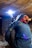 A welder wearing protective gear is working in a confined space, possibly under a structure. The bright arc of the welding torch illuminates the area, casting a blue and white light. Sparks fly as the metal is being welded, and the worker is crouched down, focusing intently on the task.