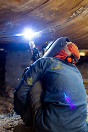 A welder wearing protective gear is working in a confined space, possibly under a structure. The bright arc of the welding torch illuminates the area, casting a blue and white light. Sparks fly as the metal is being welded, and the worker is crouched down, focusing intently on the task.