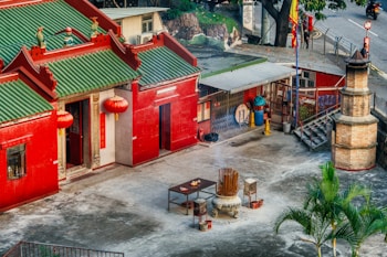 A traditional Chinese building with a distinctive green tiled roof and bright red walls is visible. The structure features ornate architectural details, including red lanterns hanging at the entrance. The courtyard is open and contains a large incense burner surrounded by smaller offerings. A palm tree is visible at the bottom right, and in the background, there are additional structures with red tiled walls and a flagged sidewalk.