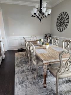A dining room featuring a wooden dining table with six ornate chairs. The table is set with decorative placemats and a centerpiece consisting of small plants. A large, black chandelier with white shades hangs above the table. On the wall is a circular decorative piece with intricate metalwork. The room has a neutral color palette with white and light gray walls, complemented by a dark hardwood floor and an area rug under the table.