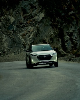 A white SUV travels along a winding mountain road, surrounded by rugged cliffs and rocky terrain. The natural landscape is dramatic and contrasts with the smooth, modern vehicle. The atmosphere is serene, with overcast lighting casting soft shadows.