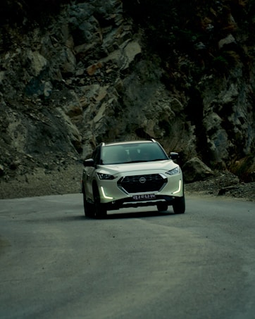 A white SUV travels along a winding mountain road, surrounded by rugged cliffs and rocky terrain. The natural landscape is dramatic and contrasts with the smooth, modern vehicle. The atmosphere is serene, with overcast lighting casting soft shadows.