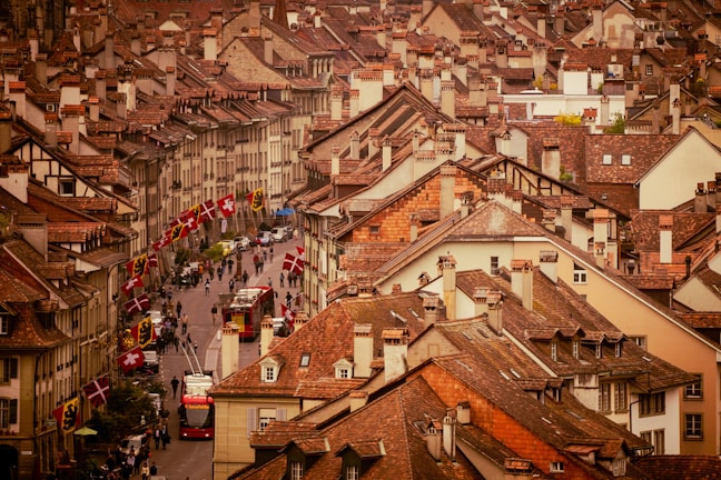 Rooftops of a densely packed city area with uniform architectural style featuring reddish-brown tiles. A street lined with Swiss flags runs through the center, populated by pedestrians and a red bus. The scene is a blend of historic charm with a lively urban atmosphere.