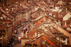 Rooftops of a densely packed city area with uniform architectural style featuring reddish-brown tiles. A street lined with Swiss flags runs through the center, populated by pedestrians and a red bus. The scene is a blend of historic charm with a lively urban atmosphere.