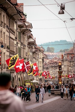 A bustling street lined with traditional European-style buildings, adorned with various national flags. People are walking, socializing, and enjoying the day in the vibrant and historic surroundings. A prominent statue stands on the right, adding to the cultural atmosphere.