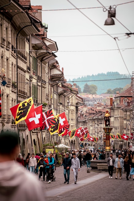 A bustling street lined with traditional European-style buildings, adorned with various national flags. People are walking, socializing, and enjoying the day in the vibrant and historic surroundings. A prominent statue stands on the right, adding to the cultural atmosphere.