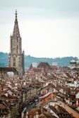 A historic cityscape featuring a tall, ornate church tower rising above rows of traditional European houses with sloped roofs. Swiss flags hang along the narrow street, and trees and hills can be seen in the distance under an overcast sky.