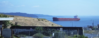 A large cargo ship is sailing on a calm body of blue water. In the foreground, a grassy hillside descends to the shore. There is a sign for Tacoma Narrows Federal Credit Union, along with some fencing and other industrial structures. The distant background features a low, distant coastline covered with trees.