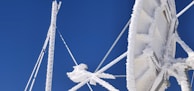 A large satellite dish covered in ice and snow is set against a clear blue sky. The ice creates intricate patterns on the surface and structure of the dish, highlighting the cold weather conditions. The aerials and supporting structures are also encrusted with frost.
