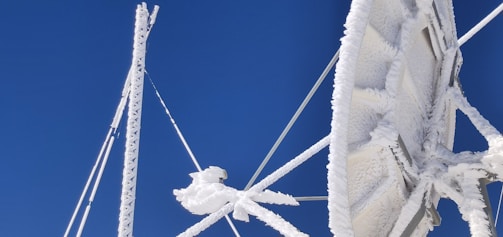 A large satellite dish covered in ice and snow is set against a clear blue sky. The ice creates intricate patterns on the surface and structure of the dish, highlighting the cold weather conditions. The aerials and supporting structures are also encrusted with frost.