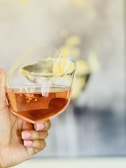 A hand with manicured nails painted in light pink holds a tilted wine glass filled with a rosé liquid. The background is softly blurred with warm, neutral tones and hints of soft yellow details.