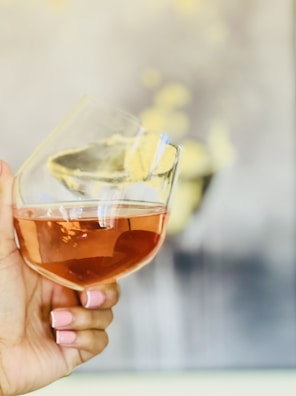 A hand with manicured nails painted in light pink holds a tilted wine glass filled with a rosé liquid. The background is softly blurred with warm, neutral tones and hints of soft yellow details.
