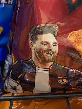 A close-up of a snack packet featuring a stylized illustration of a person with a modern haircut and beard. The name 'Messi' is visible near the figure, along with a soccer-themed graphic. The background of the packet is predominantly a dark red with portions of a bright orange snack visible.