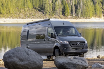 A grey van is parked near a scenic lake. Large rocks are situated in the foreground, and the background consists of a forest with tall evergreen trees. The water in the lake is calm with reflections of the trees.