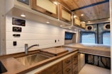 The interior of a camper van features a compact kitchen area with a sink, wooden countertop, and multiple drawers. There are overhead cabinets with warm lighting, along with wood-paneled ceiling and white wall accents. In the background, a cushioned bed is positioned at the rear of the van.