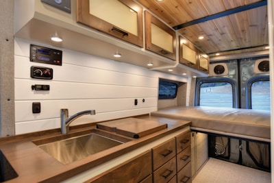 The interior of a camper van features a compact kitchen area with a sink, wooden countertop, and multiple drawers. There are overhead cabinets with warm lighting, along with wood-paneled ceiling and white wall accents. In the background, a cushioned bed is positioned at the rear of the van.