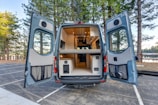 A camper van with its rear doors open, revealing a well-organized and compact interior setup. The inside is designed for functionality and comfort, featuring a kitchenette with cabinets and a wooden ceiling with recessed lighting. The van is situated in a serene outdoor setting, surrounded by tall pine trees and a glimpse of a lake in the background.