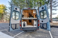 A camper van with its rear doors open, revealing a well-organized and compact interior setup. The inside is designed for functionality and comfort, featuring a kitchenette with cabinets and a wooden ceiling with recessed lighting. The van is situated in a serene outdoor setting, surrounded by tall pine trees and a glimpse of a lake in the background.