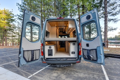 A camper van with its rear doors open, revealing a well-organized and compact interior setup. The inside is designed for functionality and comfort, featuring a kitchenette with cabinets and a wooden ceiling with recessed lighting. The van is situated in a serene outdoor setting, surrounded by tall pine trees and a glimpse of a lake in the background.