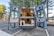 A camper van with its rear doors open, revealing a well-organized and compact interior setup. The inside is designed for functionality and comfort, featuring a kitchenette with cabinets and a wooden ceiling with recessed lighting. The van is situated in a serene outdoor setting, surrounded by tall pine trees and a glimpse of a lake in the background.